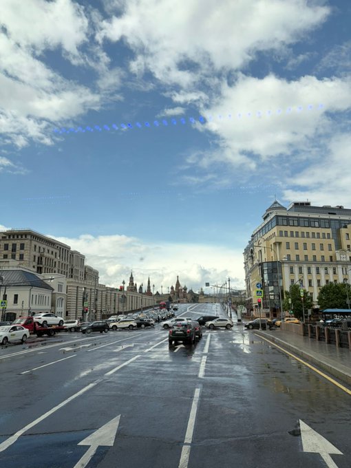 A view from inside a vehicle driving down a wet city street in Moscow, with the Kremlin visible in the distance under a partly cloudy sky.