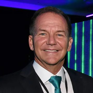 A smiling man in a dark suit and teal tie appears at what looks like a corporate event, with blue and green lit panels in the background. The image accompanies news about Paul Tudor Jones' Tudor Investment Corporation taking a $22.9 million position in BlackRock's spot Bitcoin ETF, suggesting the man pictured is the billionaire hedge fund manager himself.
