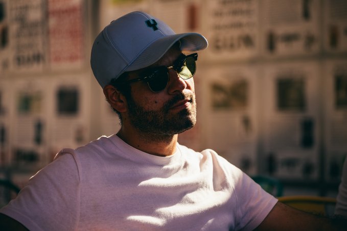 A man wearing a light blue cap, dark sunglasses, and a white t-shirt sits in front of a wall covered in newspaper clippings.