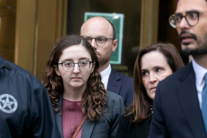 A young woman with long brown hair and round glasses, identified as former Alameda Research CEO Caroline Ellison, exits a building surrounded by security personnel and legal team members. Her serious expression reflects the gravity of her involvement in the FTX collapse, as she now faces CFTC sanctions barring her from trading futures, options, and swaps for 5 years.