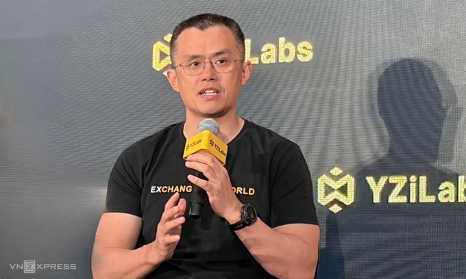 Changpeng "CZ" Zhao, co-founder of Binance, speaks into a microphone at the Bitcoin Asia 2026 conference in Hong Kong, with "YZi Labs" branding visible in the background. His black t-shirt reads "EXCHANGE THE WORLD," aligning with the Vietnamese caption describing his remarks that Bitcoin could surpass gold in importance and market capitalization in the next bull cycle, as gold's market cap is still about 10 times larger than Bitcoin's.