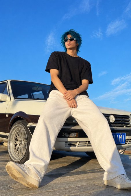 A young man with blue hair and sunglasses poses confidently in a black t-shirt and white pants, sitting on the front of a vintage Volkswagen against a clear blue sky.