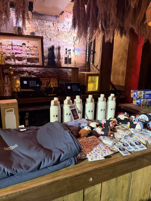 A display table at a trendy, dimly lit venue showcases "Fomo" merchandise including white water bottles, plush toys, stickers, and branded apparel. The caption aligns with the post, confirming that the Fomo merch on display was a highlight of the event.
