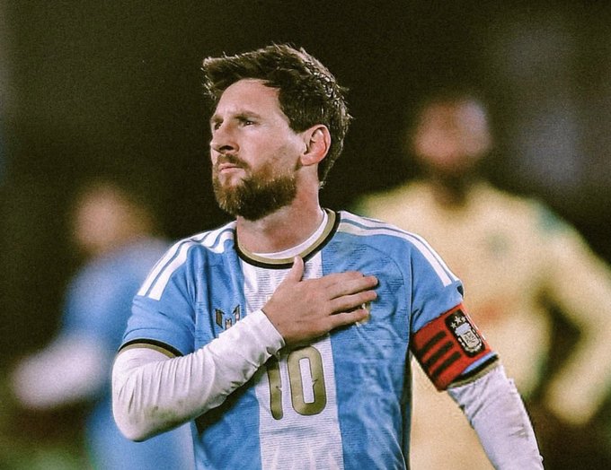 Lionel Messi wears the #10 Argentina jersey and captain's armband, with his hand over his heart in a solemn pose.