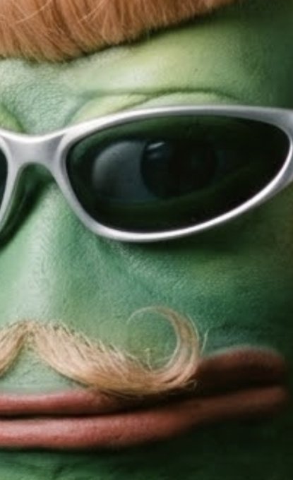 Close-up of a person wearing green face paint, dark sunglasses, a curly light-brown mustache, and reddish hair.