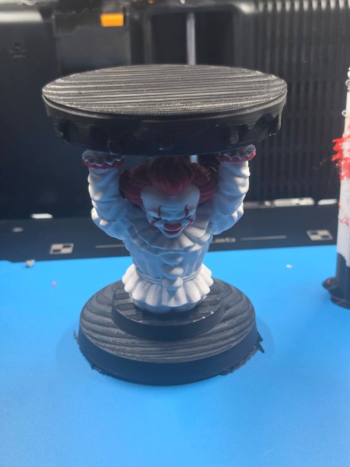 A 3D printed figurine of Pennywise the clown holding up a black sewer cover, positioned on a black base in front of what appears to be a 3D printer.