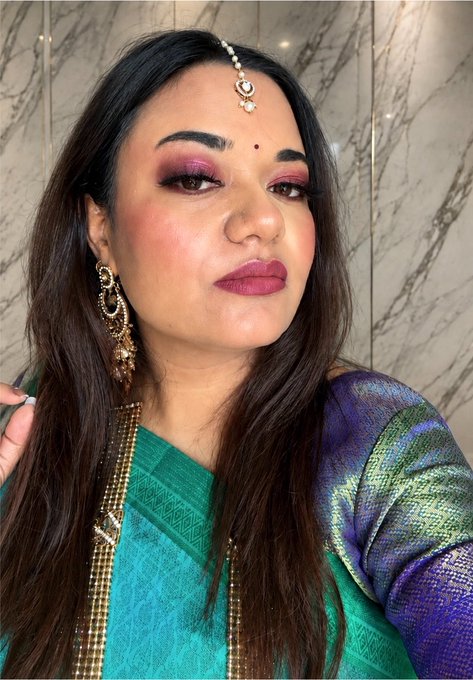 A selfie-style photo of a woman wearing traditional Indian attire including a green and blue silk saree, ornate gold jewelry, and a maang tikka headpiece, with elegant makeup featuring pink eyeshadow and lipstick.