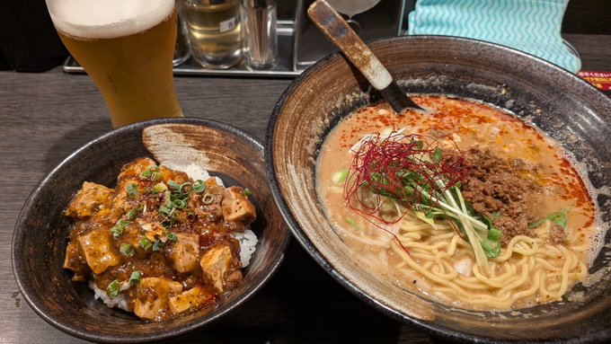A meal consisting of a bowl of spicy ramen with meat, green onions, and red garnish, alongside a bowl of rice topped with tofu in a red sauce, and a glass of beer on a wooden table.