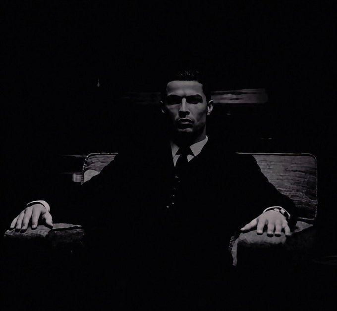 A black-and-white stylized portrait of a man in a dark suit and tie sitting in a chair, looking directly at the camera with dramatic low lighting.