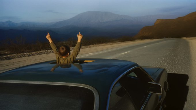 A person leans out of a car window with both arms raised in celebration as the vehicle cruises down an open mountain road, embodying the freedom and exploration that the evolving 150-piece "Unfinished Past" collection by @Dalos invites collectors to experience.
