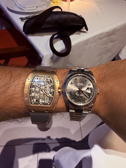 A wrist displays a gold Richard Mille watch alongside a steel Rolex Datejust II, while the background shows a handbag dropped on a white tablecloth—serving as the ultimate flex against anyone being "mogged."
