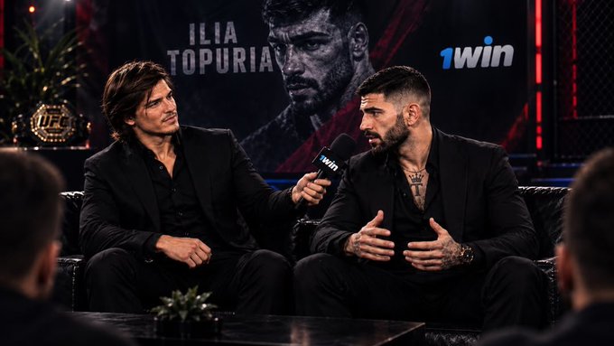 A scene featuring two men in black suits seated in leather chairs during what appears to be an interview setting, with a "1win" logo and an "ILIA TOPURIA" UFC promotional backdrop behind them.