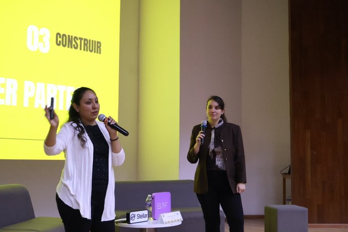 Two women speak at a conference in front of a yellow slide reading '03 CONSTRUIR', with sponsor logos visible on a small table.