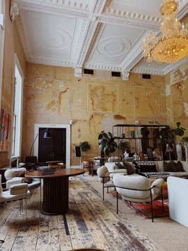 An elegant, high-ceilinged lounge featuring ornate plasterwork, a gold chandelier, and a curated mix of contemporary furniture atop a patterned rug. The refined, eclectic interior complements the architectural and design aesthetic associated with House of Sol by SuperteamUK and archill.