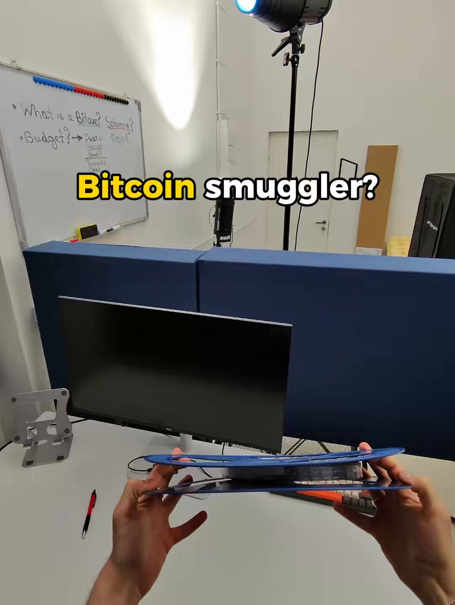 The image shows a person holding two blue computer monitors, with one of them being a flat screen monitor and the other a smaller laptop-like device. The person appears to be using these devices for work or entertainment purposes. In the background, there is a whiteboard that has some writing on it, possibly containing instructions or ideas related to Bitcoin mining or other topics.