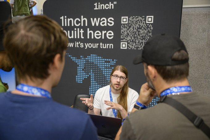 Three people wearing lanyards converse at a booth with '1inch was built here' text and a QR code visible on the display board behind them.