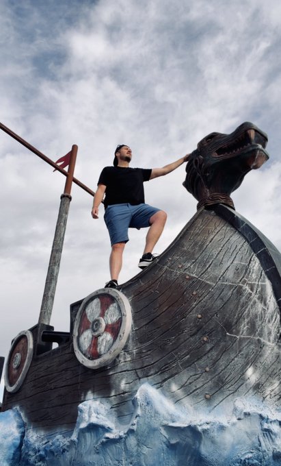 A man in a black t-shirt and blue shorts climbs on a large wooden Viking ship sculpture with a dragon prow, posed as if on stormy seas.