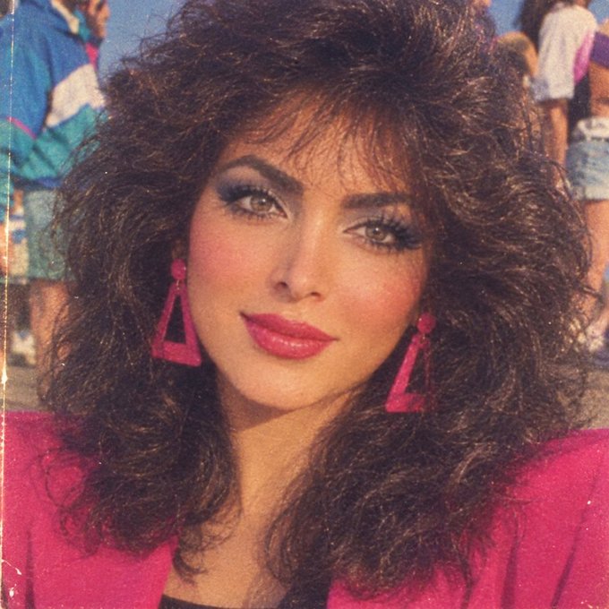 A portrait of a woman with voluminous, teased dark hair and dramatic 80s-style makeup, wearing large triangular pink earrings and a red top.
