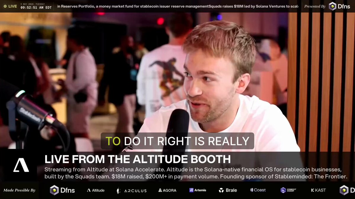 The image shows a man sitting at a microphone during an event or gathering, likely a live stream. The background of the image features a crowd of people, indicating that this could be a social media event or a public speaking engagement. The main focus is on the man and his interaction with the camera, as he appears to be talking into it.