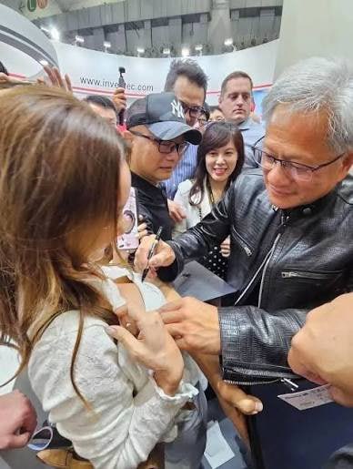 An older man in a black leather jacket signs an autograph for a fan at what appears to be a convention, with a 'www.invoice...tec.com' banner visible in the background.