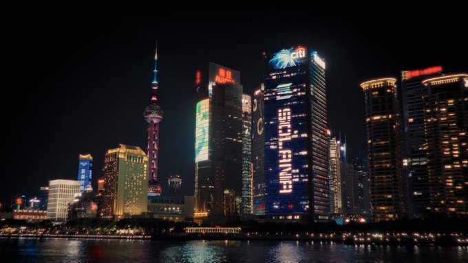 A nighttime cityscape of Shanghai's Pudong skyline along the Huangpu River showcases illuminated skyscrapers, including the Oriental Pearl Tower and a Citi building displaying the text "Solana" in vertical blue lights. The image visually represents the comparison between Ethereum and Solana, with Solana's prominent, eye-catching signage along the waterfront symbolizing its active, attention-seeking market presence as described in the post.