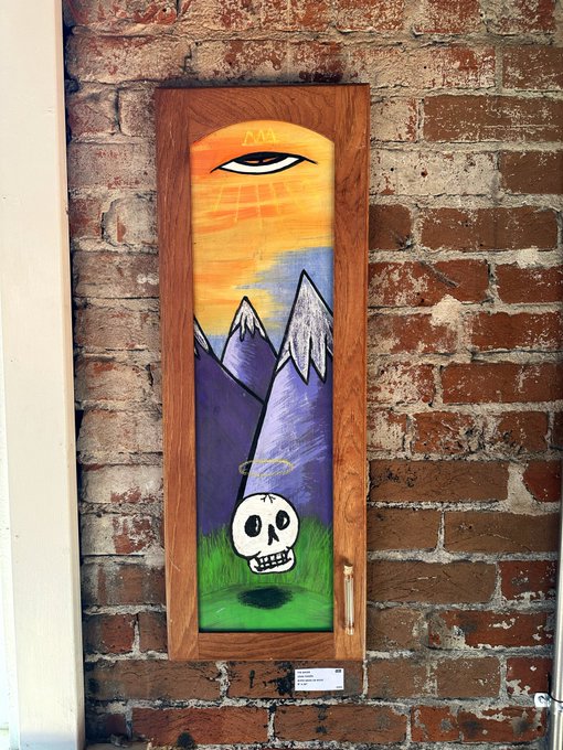 A painted wooden door mounted on a brick wall, featuring artwork of a sun, mountains, green grass, and a floating skull with a peace symbol above it.