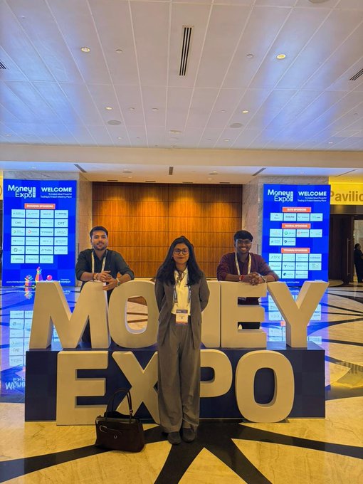 Three people pose behind a large gold "MONEY EXPO" sign at a conference, with digital displays showing "Money Expo WELCOME" in the background.
