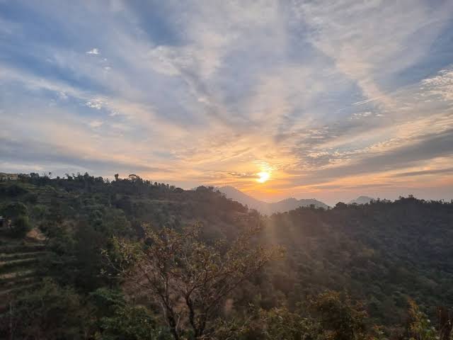 A scenic photograph captures a sunset over a hilly, forested landscape with dramatic clouds filling the sky and sunlight filtering through the trees.