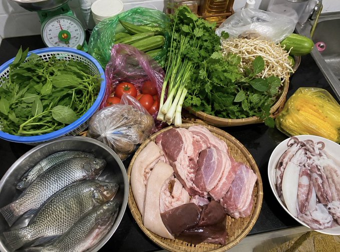 A kitchen counter displays a variety of fresh ingredients including tilapia fish, pork, squid, fresh herbs (mint, cilantro, green onions), tomatoes, and bean sprouts.