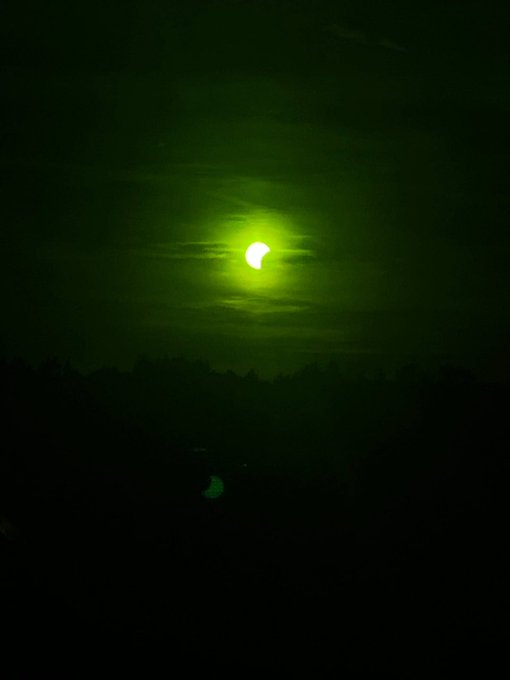 A dark photograph of a solar eclipse with a green color cast, showing the sun partially covered by the moon against a black sky above a silhouetted landscape.