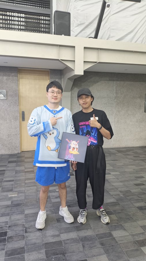 Two young men pose for a photo outdoors; one wears a blue penguin sweater and points at a framed artwork held between them.