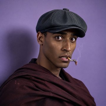 A man wearing a black cap and maroon shawl smokes a cigarette against a purple background.