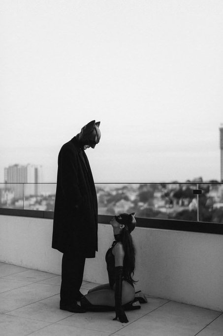 A black and white artistic photo of two figures wearing cat masks on a rooftop, with one standing in a suit and coat looking down at the other kneeling.
