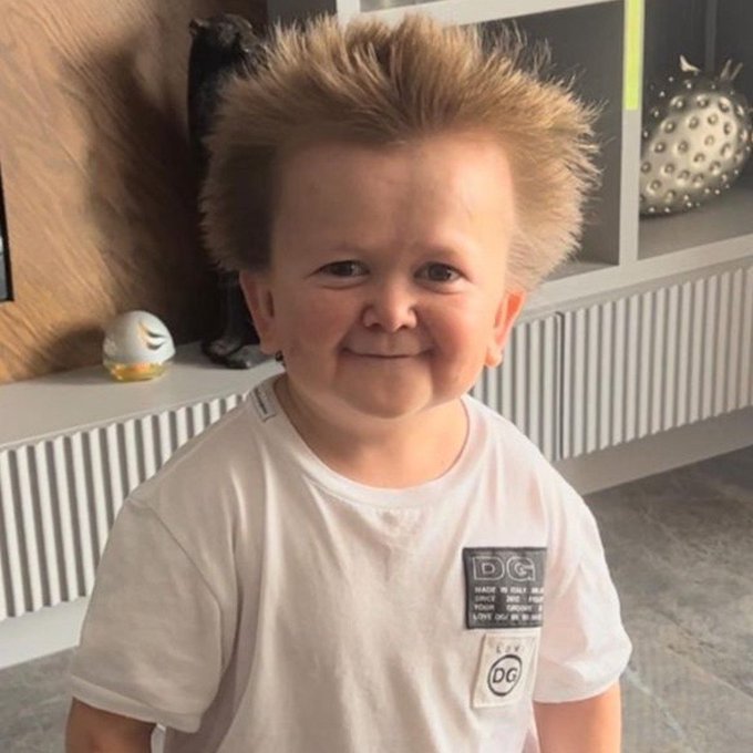 A young boy with spiked blond hair wearing a white Dolce & Gabbana (DG) t-shirt, smiling at the camera in what appears to be a living room setting.