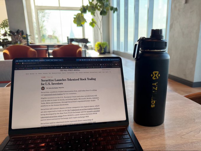 A laptop on a wooden table displays a Wall Street Journal article titled 'Securitize Launches Tokenized Stock Trading for U.S. Investors,' next to a black 'XTRIADS' branded water bottle.