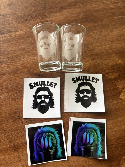 A photograph of merchandise including two glasses etched with beards, two stickers labeled '$MULLET' featuring a bearded man with sunglasses, and two colorful stickers with mullet hair designs, arranged on a wooden surface.