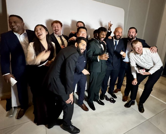 A group of approximately nine people in formal attire pose together smiling and laughing in front of a white backdrop.