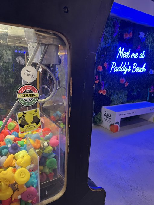 An indoor scene featuring a claw machine filled with colorful rubber ducks and figurines on the left, and a neon sign reading "Meet me at Paddy's Beach" with a "Slice" branded bench on the right.