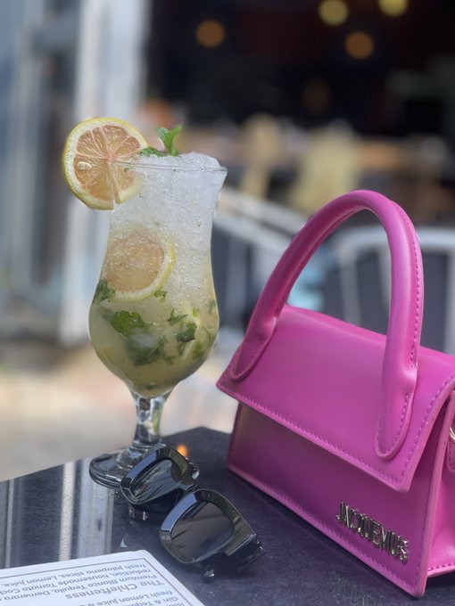 A vibrant pink Jacquemus handbag sits beside a refreshing cocktail garnished with lemon and mint, with sunglasses resting on the table—perfectly capturing a chic, leisurely vibe that complements the post's upbeat declaration of a "profitable day."