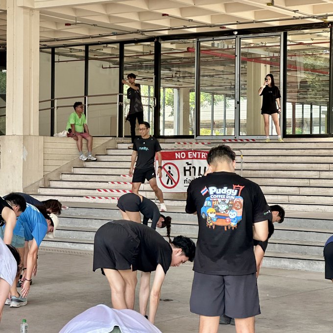 A group of people stretch and gather on the steps outside a building, with one person wearing a black 'Rudgy COFFEE RUN' t-shirt and a 'NO ENTRY' sign visible in the background.
