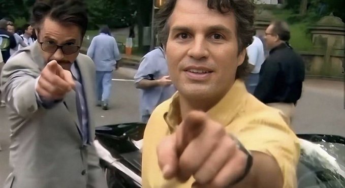 Two men, one in a yellow shirt and another in a beige blazer and glasses, both point directly at the camera with intense expressions on a crowded city street. The aggressive, confident pointing gesture paired with the post "this guy is about to print so hard" conveys a sense of bullish determination, suggesting imminent financial success.