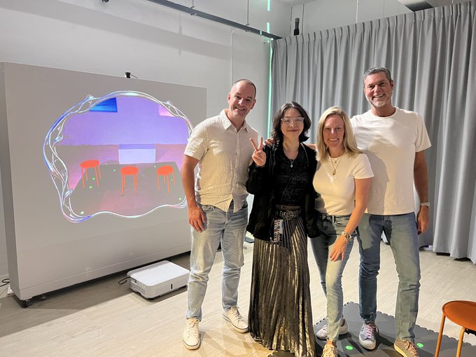 Four people posing together in a modern room with a large digital display showing an artistic rendering of a meeting space; one woman makes a peace sign.