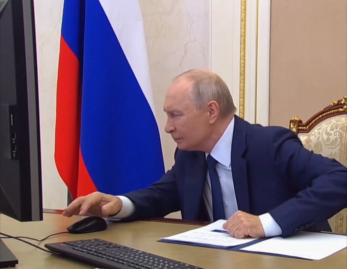 Vladimir Putin sits at a desk using a computer mouse, wearing a dark suit, with a Russian flag visible in the background.