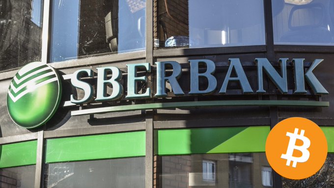 The exterior of a Sberbank branch showing its green logo and silver lettering on the building facade, with an overlaid orange Bitcoin (₿) logo in the bottom right corner.