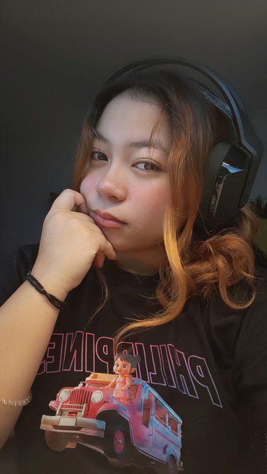 A selfie of a young woman wearing a black 'THE PRINCESS' graphic t-shirt and blue HyperX gaming headphones, resting her chin on her hand.