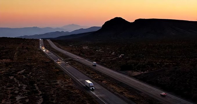 A highway at dusk with vehicles traveling through a desert landscape, framed by silhouetted mountains under a pink and orange sunset sky.