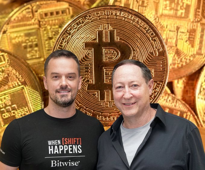 Two men pose in front of a large backdrop featuring Bitcoin coins; the younger man on the left wears a black "When [SHIFT] Happens" Bitwise t-shirt, while the older man on the right is identified as Jordi Visser (@jvisserlabs), macro strategist and guest on the podcast episode.