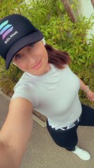 A selfie of a person wearing a black cap with a colorful logo and a white t-shirt, standing outdoors against a background of green plants.