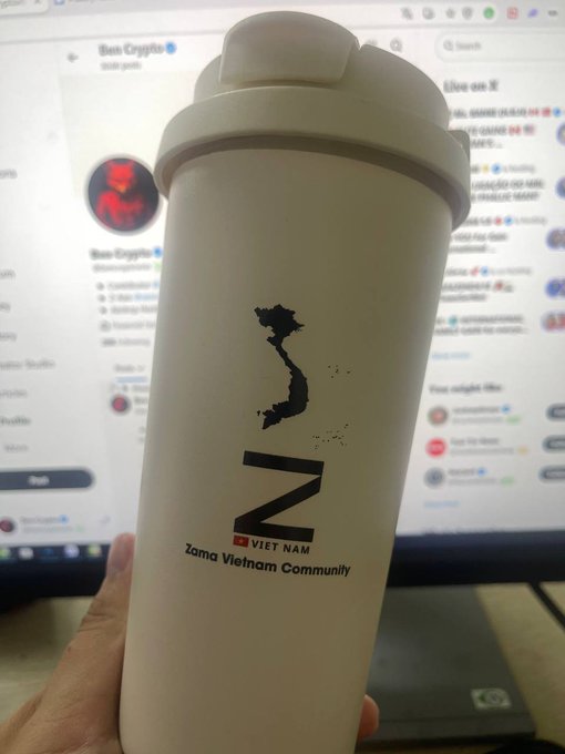A hand holds a white tumbler displaying the Vietnam map outline, a 'VN' logo, and 'Zama Vietnam Community' text in front of a computer screen showing a Twitter/X profile.