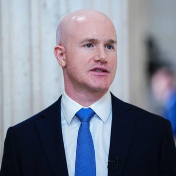 A bald man in a dark suit and blue tie appears to be speaking in an interview setting.